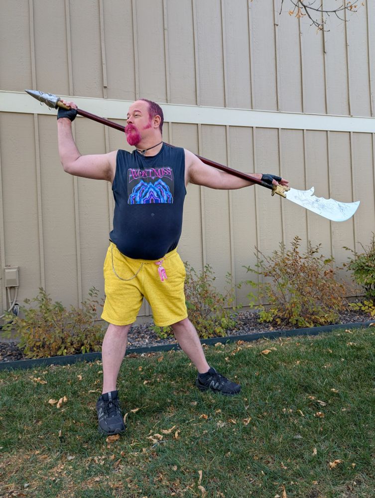 A middle-aged guy cross-playing as Mira from K-Pop Demon Hunters.  A guandao across his shoulders, magenta goatee and hair, yellow shorts, sleeveless black tee with the Huntr/x "Won't Miss" slogan.