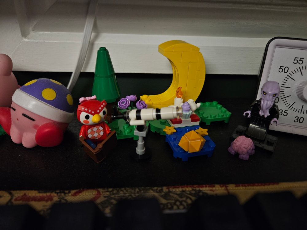 The LEGO Animal Crossing Celeste set complete and sat on my desk next to a minifigure of a mindflayer and intellect devourer. The set consists of a Celeste minifigure holding a book, telescope, and then a tea set on some grass with star fragments, a moon, and a tree and flowers.