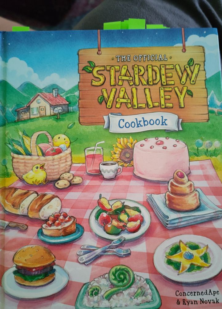 colorful painted front cover of the official stardew valley cookbook by ConcernedApe & Ryan Novak. 
different meals (like pink cake, lucky lunch, survival burger, etc.) known from the game,  on a red/white checkered table in the front. In the back are green hills and a tiny farmhouse.