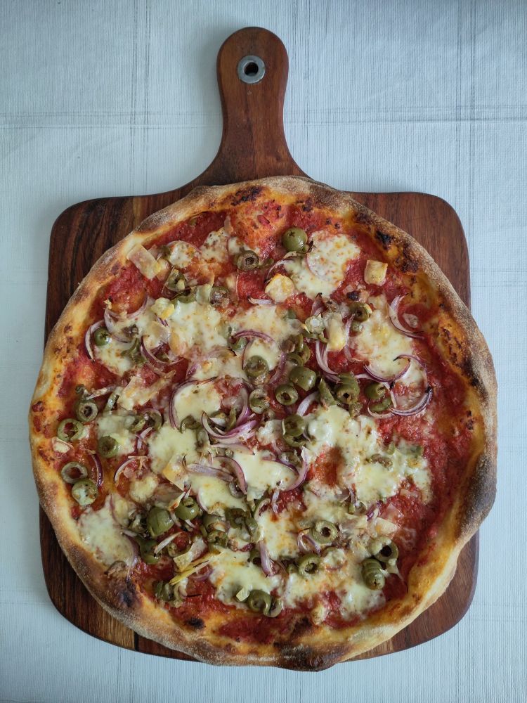 wheat sourdough pizza with a darker brown crust and tomato-basil sugo, green olives, red onions, gouda, and parmesan cheese, after baking, on a wooden board.