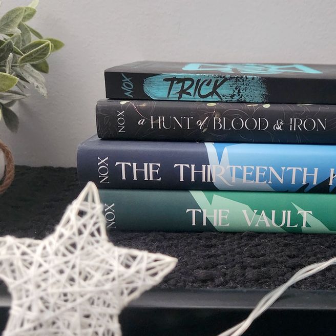 A stack of books by Cara Nox, from top to bottom: Trick, a black book with teal paint grunge design; A Hunt of Blood & Iron, a black book with hints of plants and a faded gold pattern; The Thirteenth Key, a book with shades of blue; and The Vault, a book with shades of green. The books sit against a grayish wall with a plant cut off along the edge of the photo and a woven white star light, looking out of focus, in the foreground.