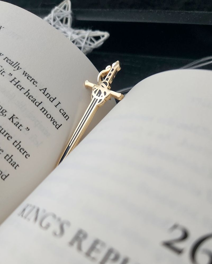 An open copy of the book "The Thirteenth Key" by Cara Nox with a rapier pin between the pages as it rests on a black tray with woven white star lights in the background.