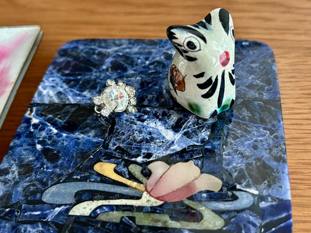 藍色の石に桃色の花と流水のような曲線が描かれた象嵌のコースター。その上に白い宝石とシルバーが複数ついたイヤリング1個と、白地に黒い線や赤、緑などで素朴な着彩がされた猫らしき生き物の小さな置き物（焼き物）が置かれている。