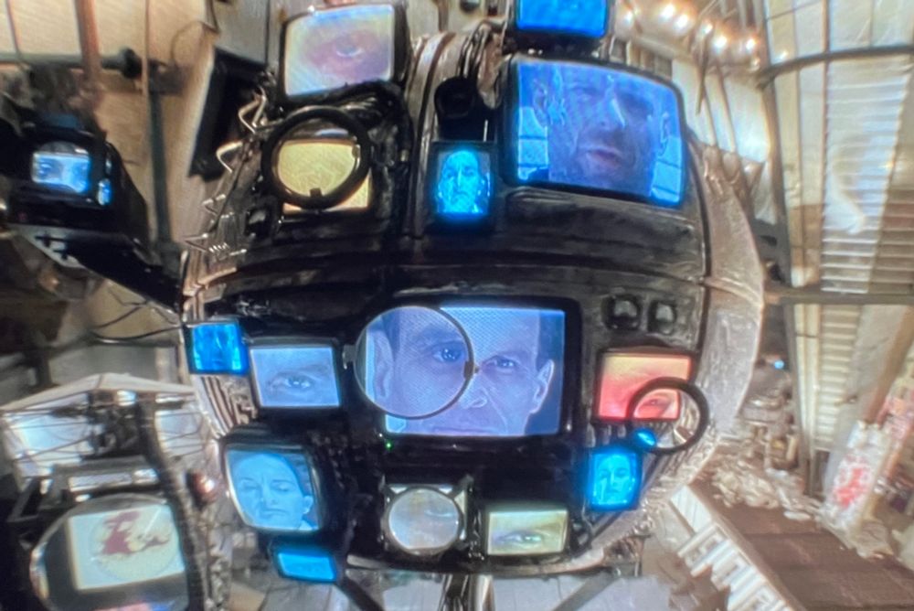 A still from Twelve Monkeys (1995) shows approximately 15 screens of varying sizes, mounted onto a stand, monitoring the viewer. The screens show different parts of people‘s faces. Some are super close—just the eyeball—others show a whole face.