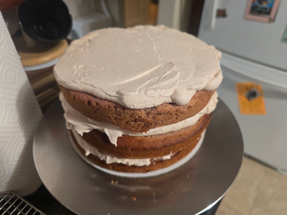 A three tier cake on a turntable with whipped cream spread on top and on between the layers