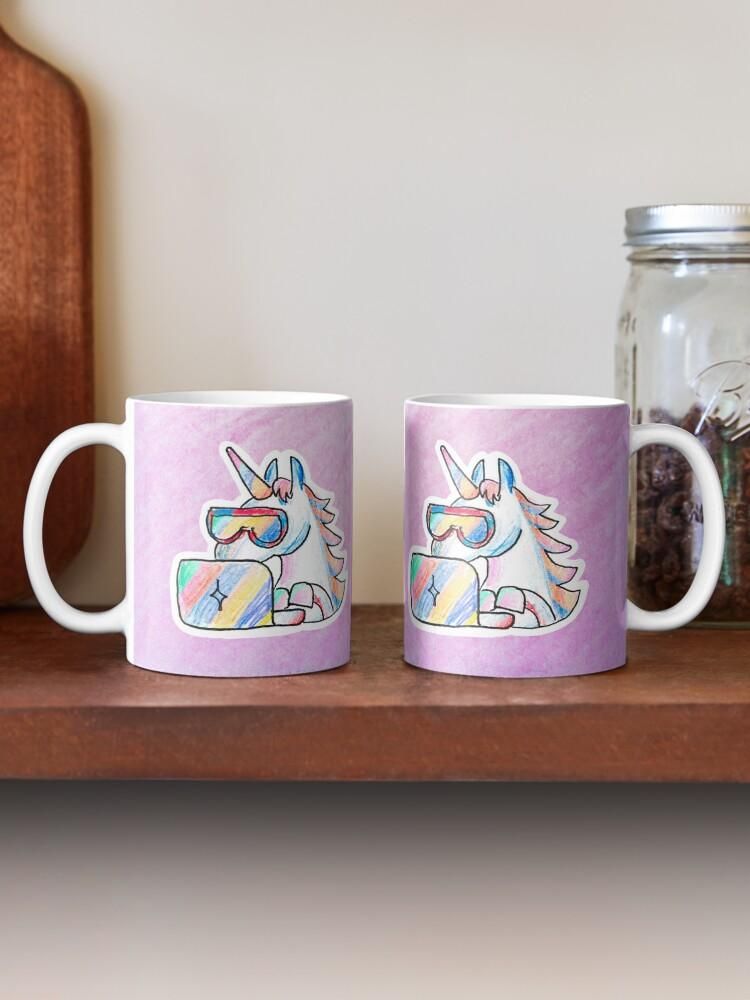 Product photo of a mug with a colorful hand-drawn unicorn wearing cool googles and typing into a notebook. The background of the artwork is rose/pink.