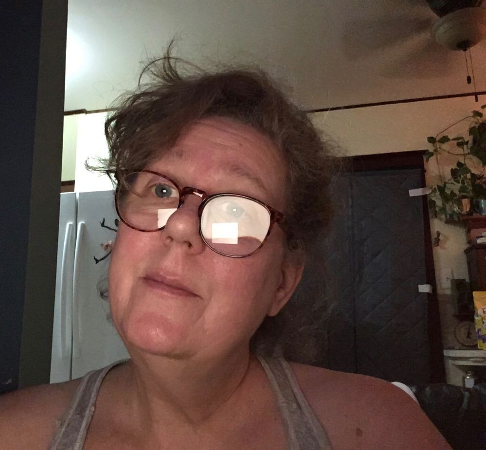 Middle aged woman with hair messily pulled back. Sitting in a moderately lit kitchen. No frills. No makeup. Unpolished.