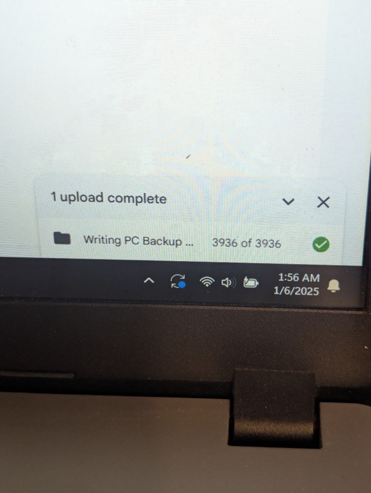 Photo of the lower right-hand corner of a laptop screen with a Google drive notification of 1 upload complete of writing pc backup... 3936 of 3936 files with a green check mark. In the corner there are generic system notifications of restart available, wifi connected, volume low, battery charging, and 1:56am 1/6/2025
