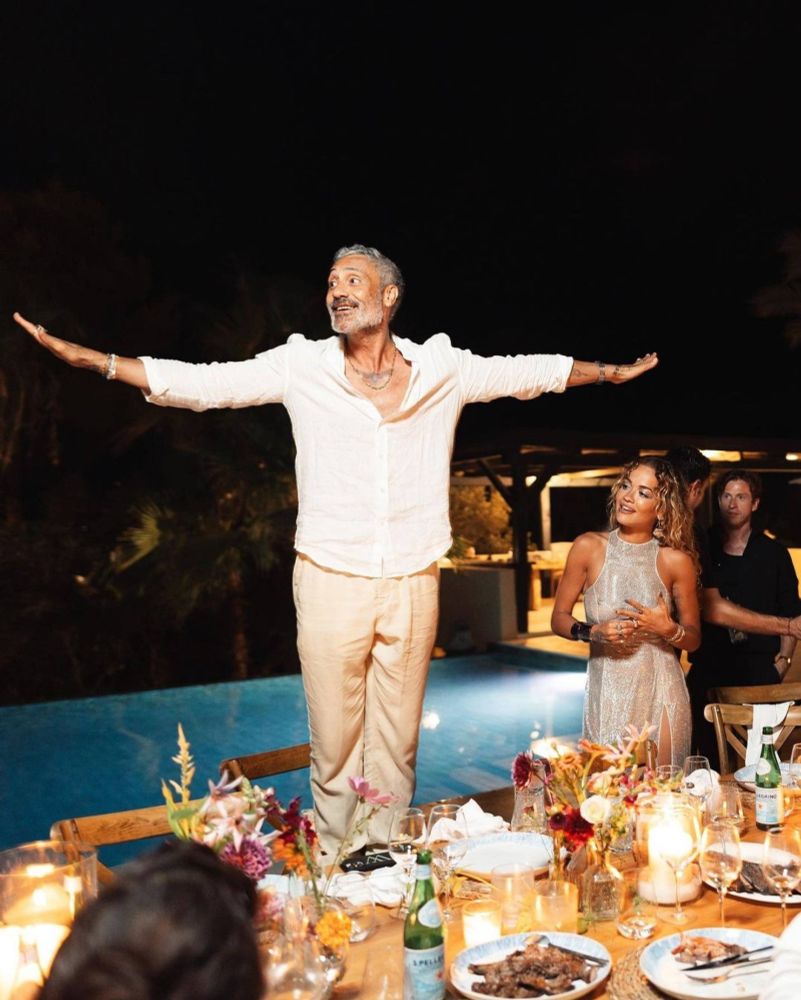 Photo of Taika at his birthday party. He is standing on a chair behind a laid table. His arms are stretched wide and he's smiling to the side like he wants to fly off. He's wearing light brown pants and a white shirt that is open at the neck so you can see the hawk tattoo (yum). Rita is on the ground on his right and looking up at him. It's night, and there's a lit swimming pool in the background.