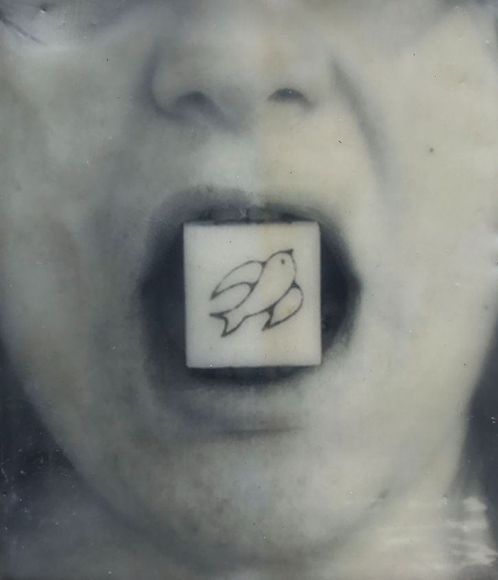 A black and white image of a woman's mouth with a wooden block between her teeth sowing a line-drawing of a bird of peace.