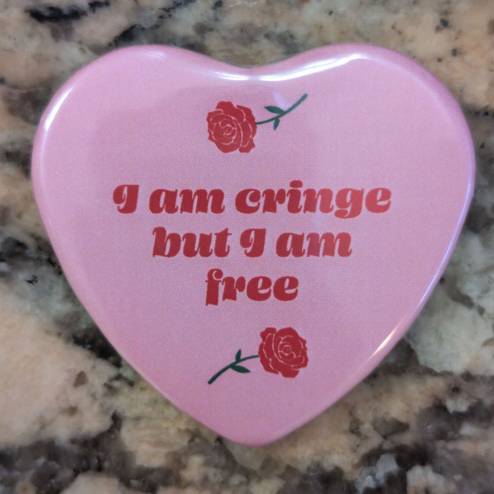 a pink heart-shaped magnet on a granite countertop. "I am cringe but I am free"is written on the magnet in red text. A rose stem appears above and below the text