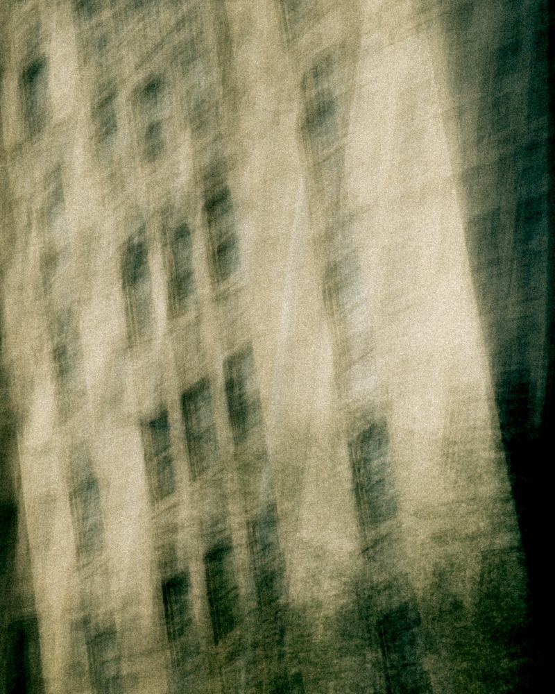 A tall building with repetitive windows appears veiled in shadow and time, its structure distorted by vertical motion. Muted tones of sepia and mossy green wash over the surface, giving the image an aged, cinematic quality—like an echo of architecture remembered in a dream.