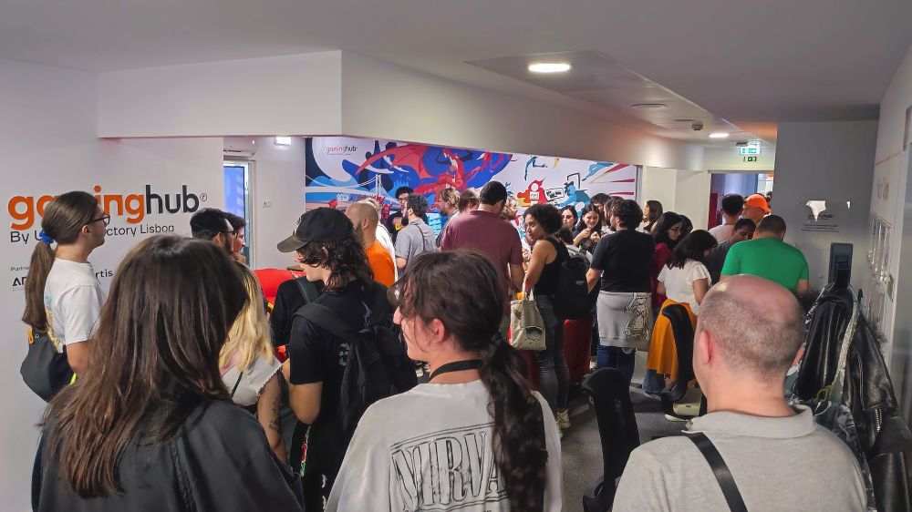 A photo of the Game Break event in Lisbon, showing a big crowd of attendees in a well-lit room. Some attendees are showing games, although most are talking with each other. A wall on the left side reads "Gaming Hub".