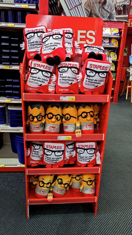 A red shelf holding various plushies at a Staples in Lisbon, designed after their own merchandising. There are two plushies being displayed on the shelves, the first being a red paper ream with a happy face wearing square glasses, and which has hands and black shoes. The writing on the plushie says it's an "A4 Paper Ream for Copy and Print" in Portuguese. The second plushie is a yellow pencil named Xavi, also with a happy face with round glasses on top of a nose sticking out. Each plushie costs 10€ to purchase.