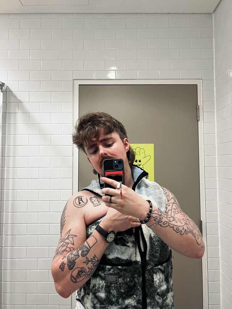 an aggressively tattooed josh in a fuzzy gilet taking photos like a himbo in the toilet 