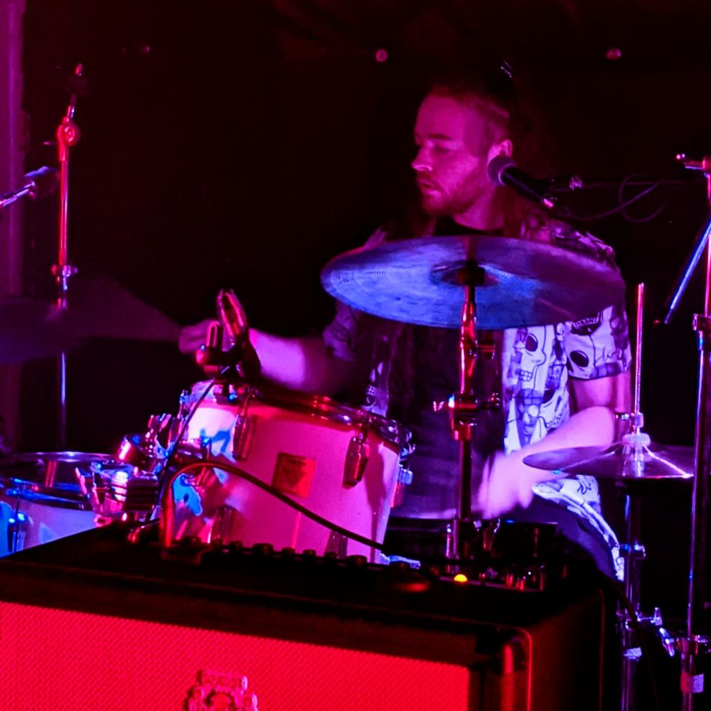 A male drummer with long red hair and a short beard, wearing a Hawaiian shirt featuring skulls.