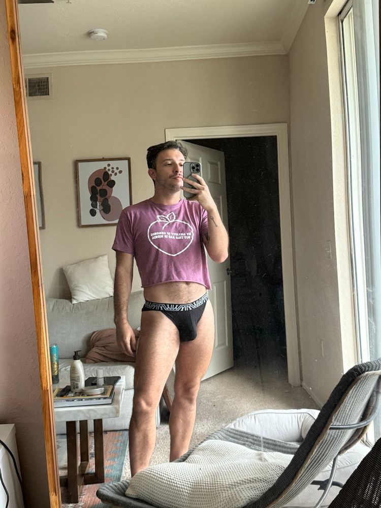 Man standing in from mirror in his black underwear, wearing an off red shirt that reads “My anxiety is chronic, but this ass is iconic”