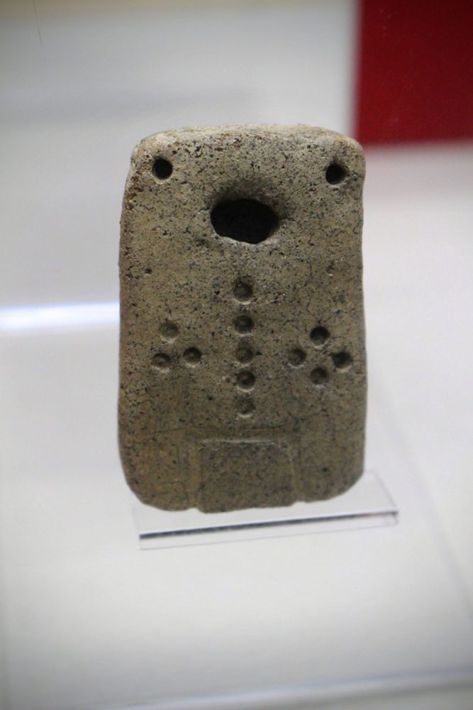 Picture of "Doban-kun" in a display case. This clay tablet figurine is decorated with a pattern of impressed dots, with a large distinctive mouth, two deeper set eyes, five dots running down the center of the figurine with three dots to the left of the centerline and 4 dots to the right.