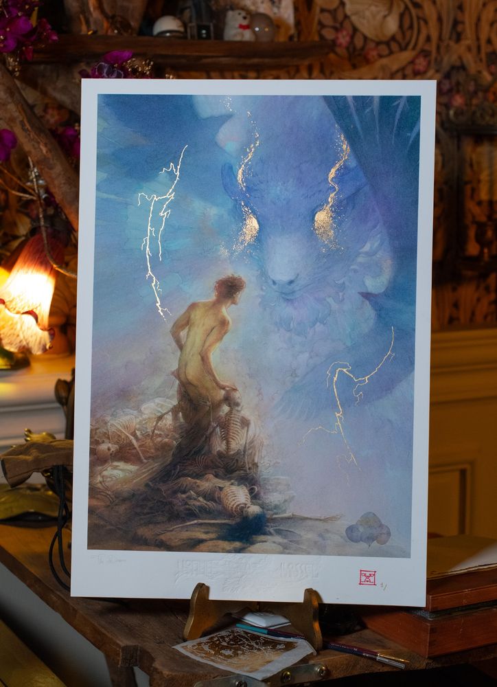 Fine art print titled The Dream, shown on a wooden stand in a warmly lit studio. The artwork depicts figure standing on a mound of skeletons, gazing up at a colossal, ethereal bison-headed deity emerging from a blue and violet dreamscape. Delicate flashes of 22K gold leaf highlight the god’s eyes and surrounding lightning.