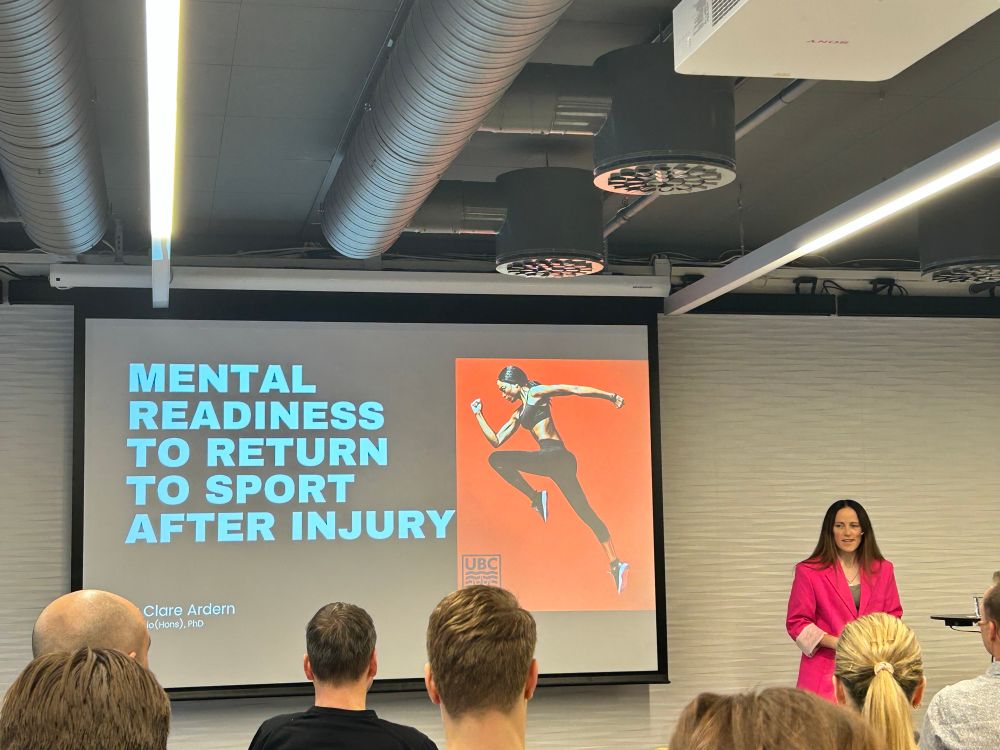dr. clare ardern stands beside her title slide which reads “mental readiness to return to sport after injury”