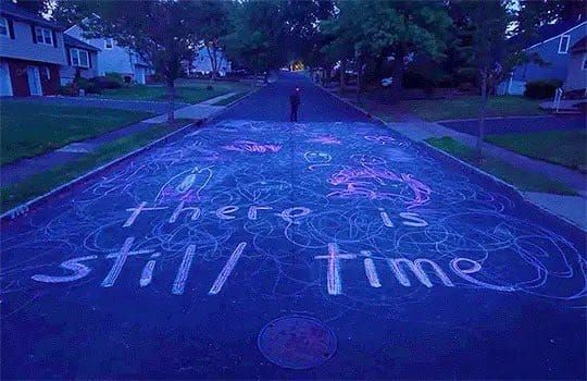 A screenshot from the movie I Saw the TV Glow.
It depicts a suburban street covered in chalk drawings. The text “there is still time” is presented front and center in the chalk.