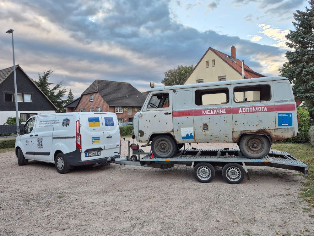 Ein Transporter mit einem Anhänger, auf dem Barwinka, ein zerschossener ukrainischer Krankenwagen steht.