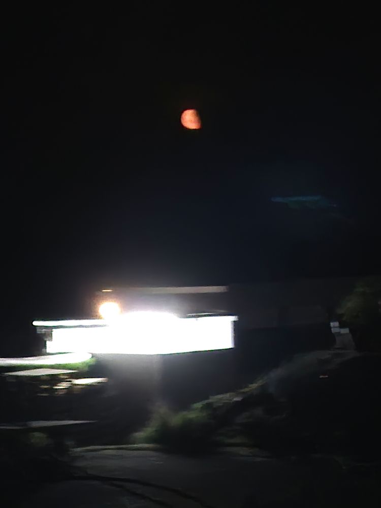 Ein grell beleuchteter Wohnwagen, darüber ist ein schöner, orange farbener Mond zu sehen.