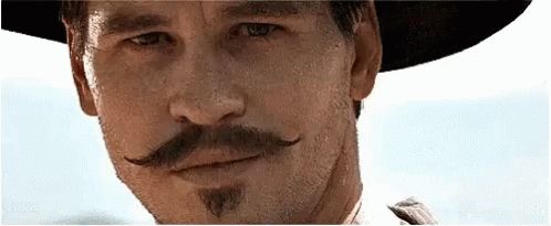 A gif of Val Kilmer playing Doc Holliday in the film Tombstone. A man looks towards the camera. He's wearing a black wide-brimmed hat and has a sharp moustache and goatee. His pallor is pasty and he looks unwell. Nevertheless, a smile plays at his lips and he's winking with his right eye.