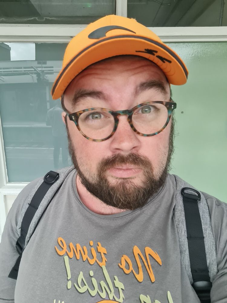 A white man with a beard, glasses and an orange hat looks at the camera