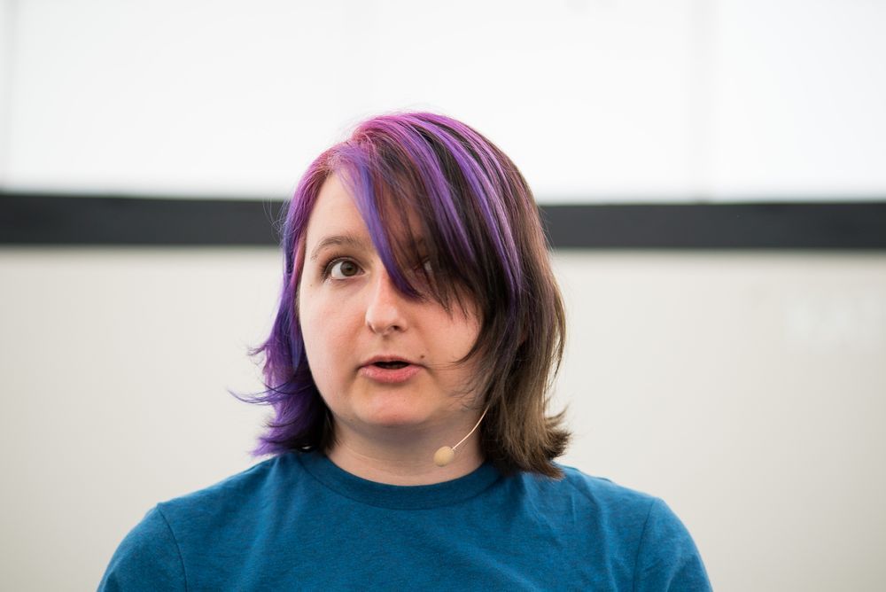 A white woman with short brown and purple hair, a blue top and a headset mic stands in front of a white background