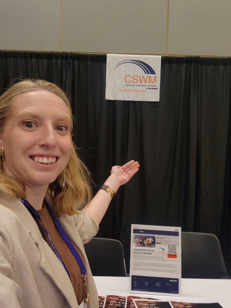 A smiling person with shoulder-length blonde hair takes a selfie while gesturing toward a booth display. Behind them is a black curtain with a white sign reading “CSWM – Commission on the Status of Women, A Commission of AEJMC.” On the table in front of the booth are flyers and a vertical sign with information and a QR code.

