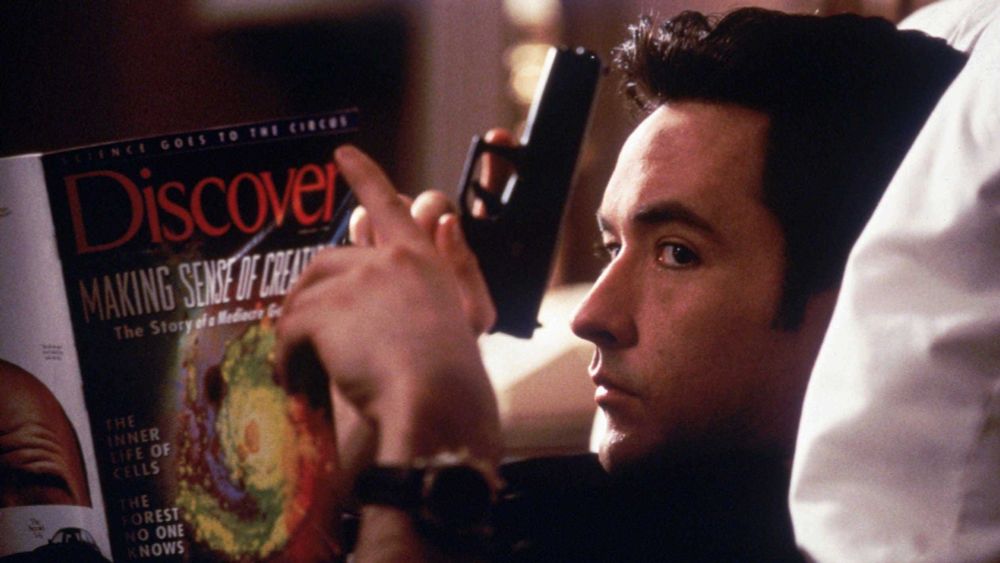 Screengrab from the movie 'Grosse Pointe Blank', featuring John Cusack holding a gun and reading 'Discover' magazine 