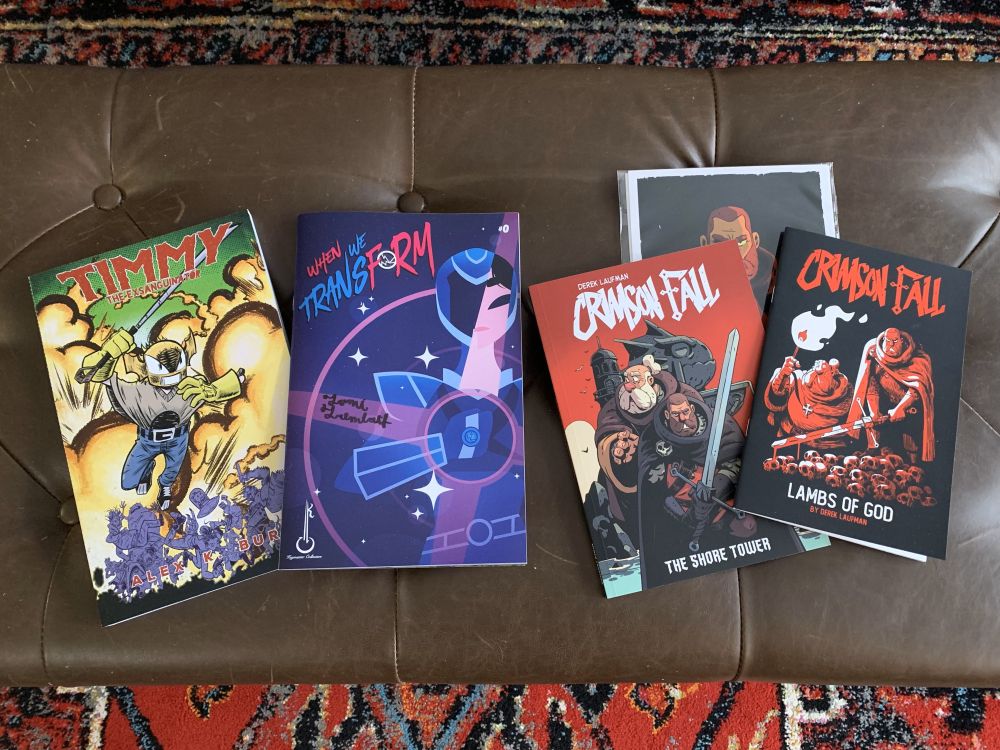 A photo of comic books lying on a leather bench. 
"Timmy the Exsanguinator" by @alexburnsart.bsky.social 
"When We Transform" by @tomitrembath.bsky.social 
"Crimson Fall" by @laufman.bsky.social 