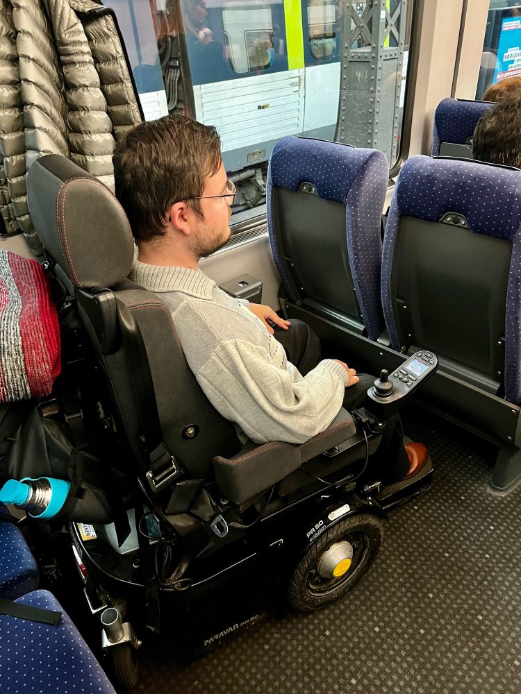 Constantin sitzt im Rollstuhl hinter einer Sitzreihe im Zug. Die Sitzreihe hat keine. Klapptisch.