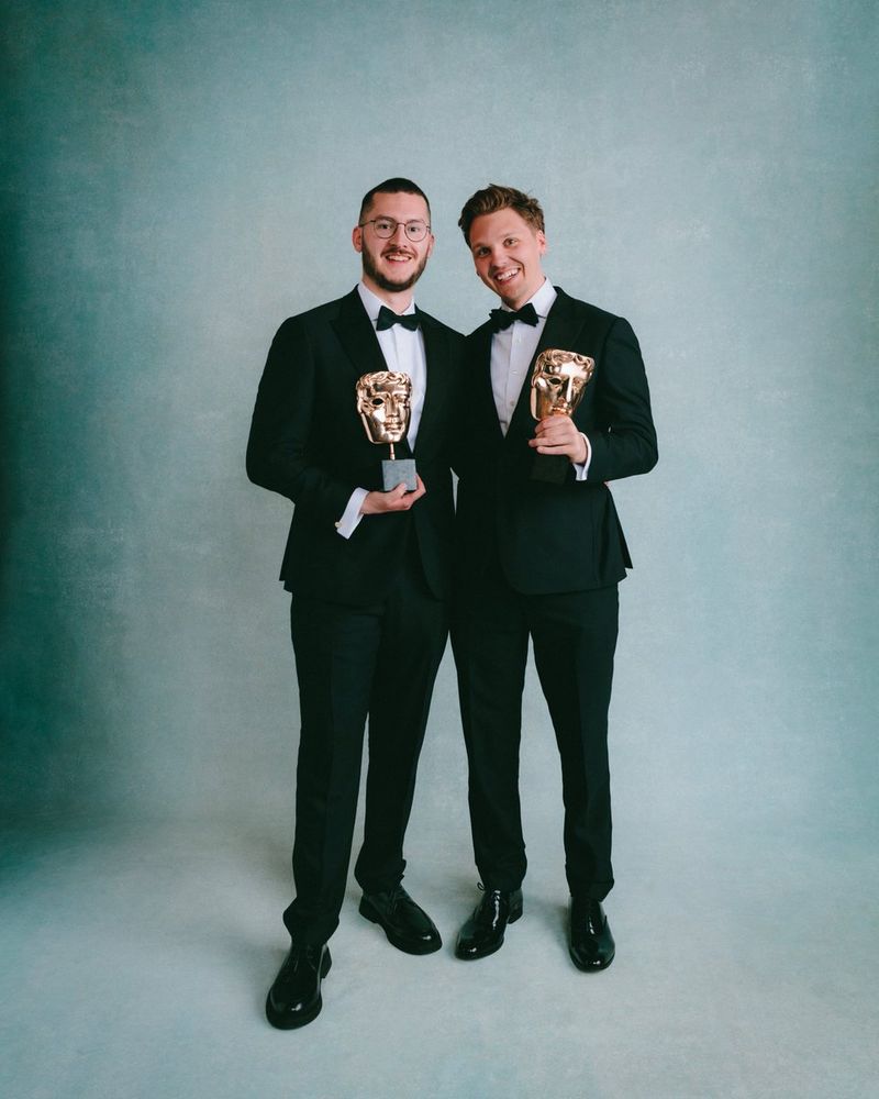 two 30 year old boys in tuxedos, holding a trophy shaped like a big gold face