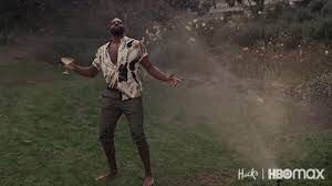 A screenshot from HBO’s Hacks. Marcus dances in the sprinklers with a drink in his hand despite his employer being fined for overconsumption of water.