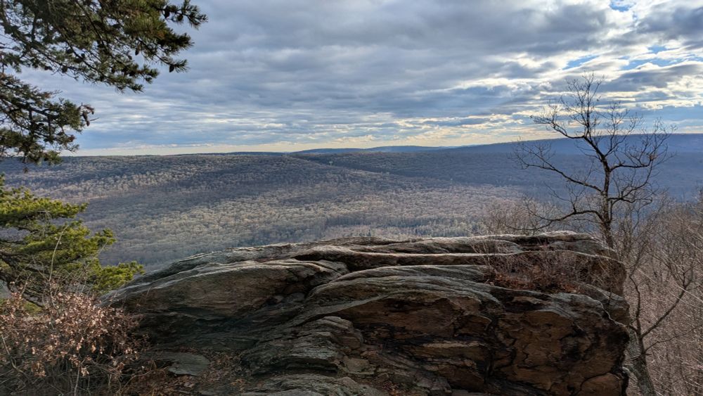 A view over a large piece of rock, looking over a lot of trees and a cloudy sky.