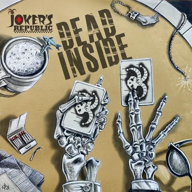 A card table with skeleton hands dealing a deck of cards with Joker's on the back. A burned out match, a lucky humans foot charm, and a drink with a shark stir stick are on the table along with a lit fuse

The table has the words Joker's Republic and Dead Inside written on it