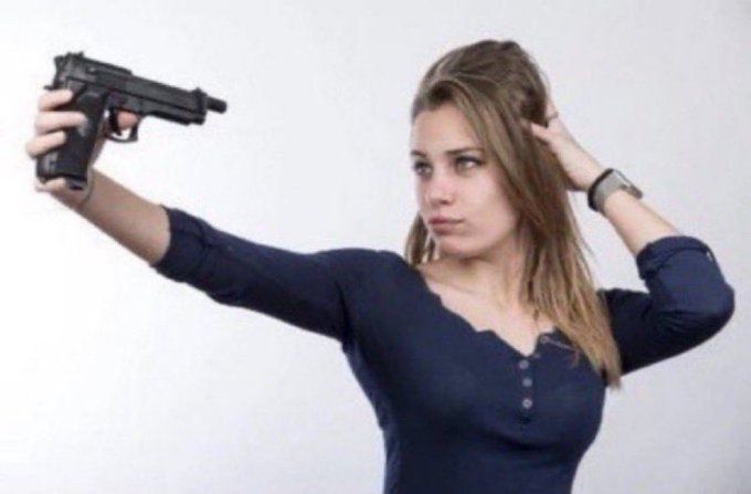 menina loira fazendo pose como se estivesse tirando uma selfie mas segurando uma arma apontada para o rosto com o braço estendido ao invés de um celular