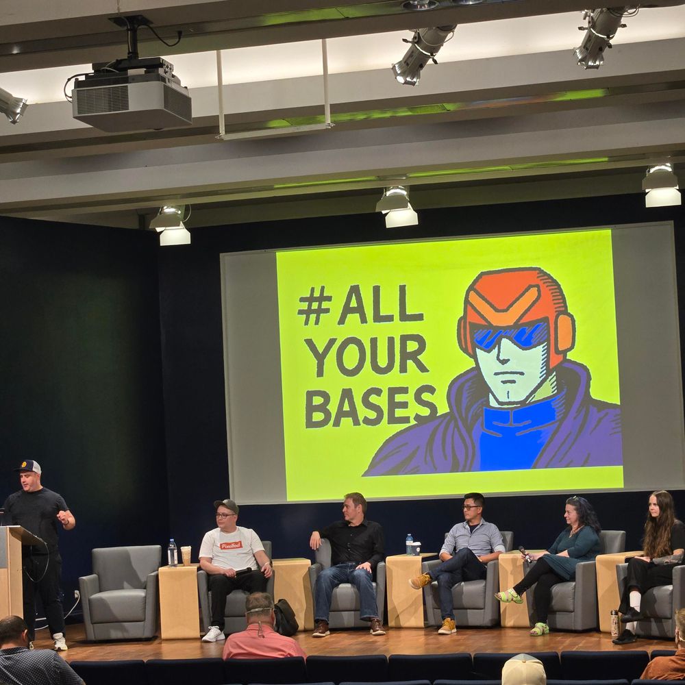 Chris Trottier on stage hosting a panel with the speakers sitting next to him. In order left to right: Dan, Ben, Julian, Johanna, and Dawn. Behind them is a giant screen that says "All Your Bases"