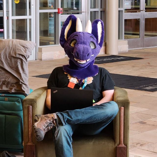 A fursuiter (head-only) sits in a chair in a hotel lobby with a laptop