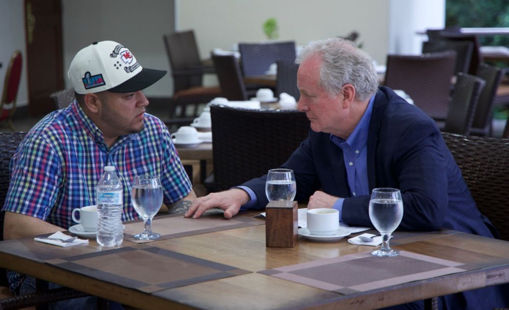 An image of Senator Van Hollen and Kilmar Abregio Garcia meeting in El Salvador. The table they are seated at is wood with plastic place mats. The room has other tables like theirs in it but no one else is in the shot. Each man has a cup of coffee, a bottle of water and a glass with water in front of them. There is no additional glass with cherries attached in this image. Senator Van Hollen is wearing a blue suit with blue shirt and no tie. He isleaning in toward Mr. Garcia with a look of concern on his face. Mr. Garcia is neatly dressed in a plaid shirt and ball cap, and looks tired, but not unduly thin or pale. 