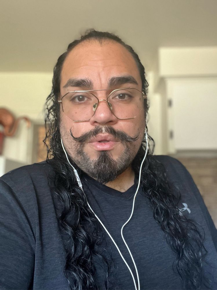 Guy with curly mustache, long curly hair, thick eyebrows, brown eyes and golden glasses wearing a grey t shirt and earphones. Blurry background