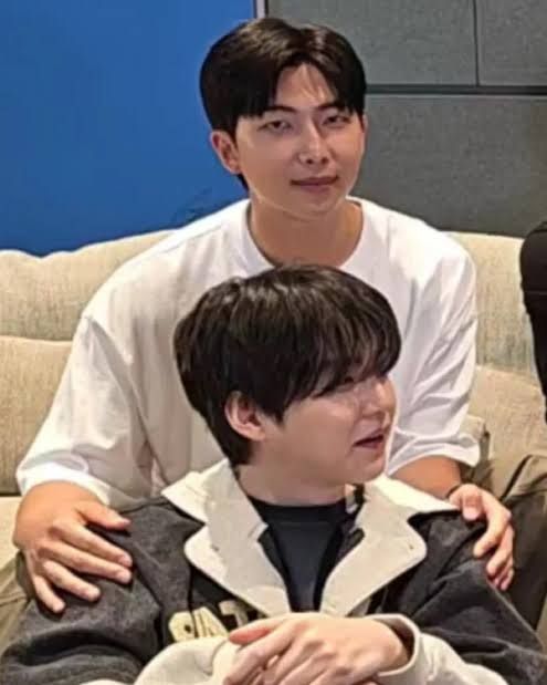A screenshot of BTS' RM and SUGA. RM, who is wearing a white T-shirt and khakis, is seating on a cream couch. He is looking at the camera.

Sitting on the floor AND BETWEEN RM's LEGS is SUGA. He is wearing a black T-shirt and a black washed jacket with cream lapels and sleeves. He is smiling while looking at someone to his left.