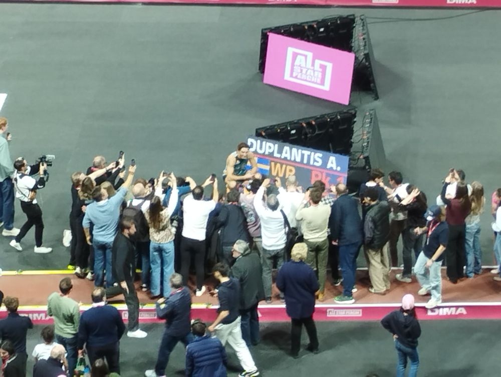 Le perchiste Armand Duplantis, entouré de photographes, est accroupi à côté d'un panneau affichant son nom et le nouveau record du monde de saut à la perche WR 6m27 