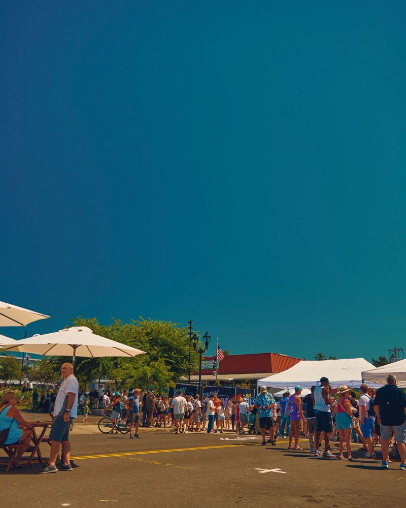 A color photo of a crowd of people meandering around white tent tops that house crafts, food, and wares under a bright, clear, blue sky.