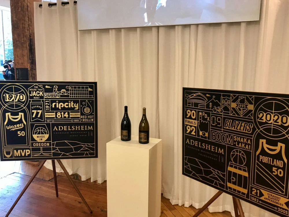 A photo showing two bottles of wine on a square pedestal, and two side by side art boards of the label concept for the Blazers’ 50th anniversary.