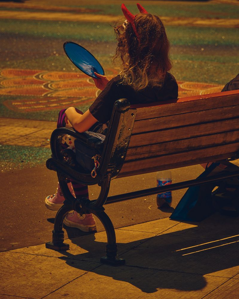 A color photo of a woman on a bench fanning herself with a frisbee. She is wearing foam horns.