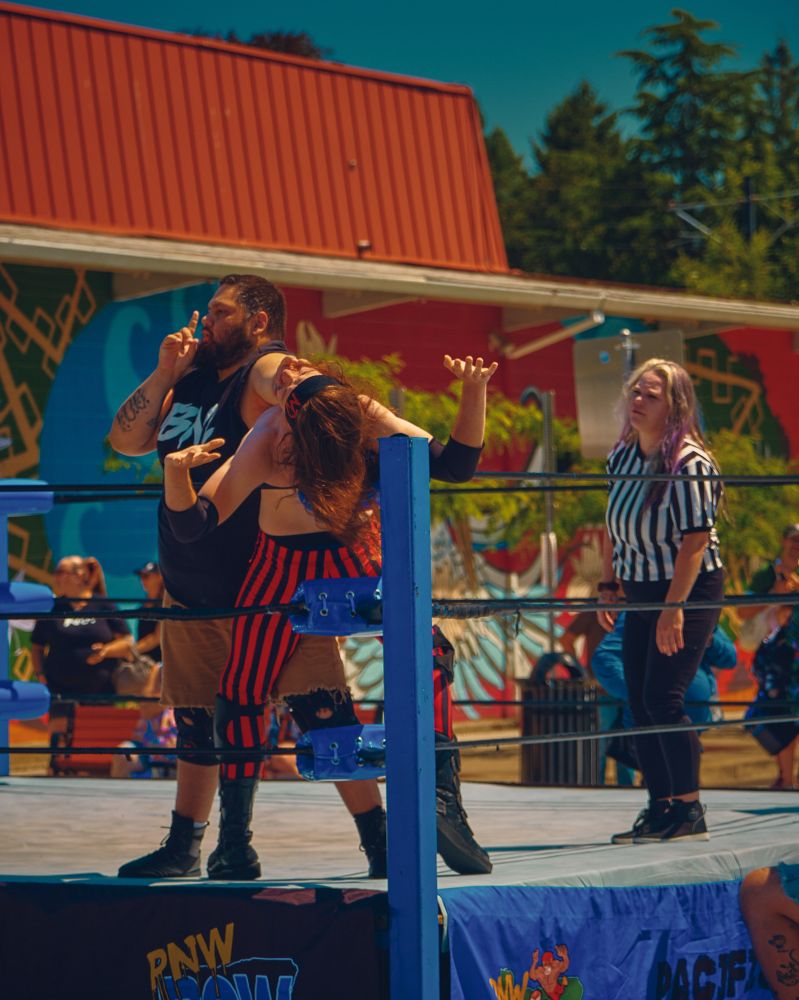 A color photo of a wrestler “shushing” the audience while holding someone against the ring post. An exhausted referee looks on in front of a colorful background.