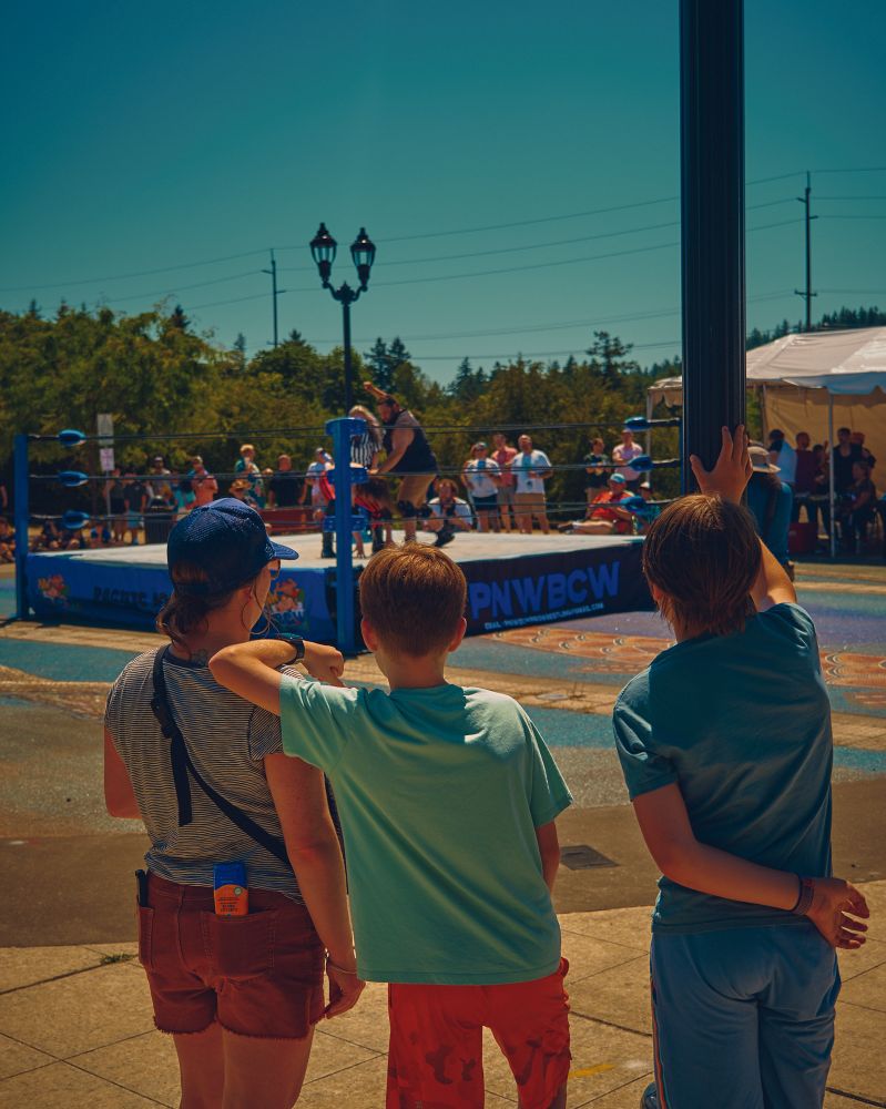 A color photo of a mom and two sons watching the wrestling match from the scorching sidewalk.